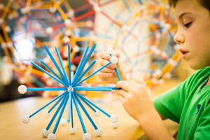 A child in a green shirt builds with Zometool. A large Zometool construction rests in the blurry background.