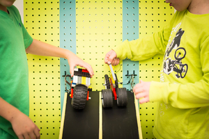 Two children are holding LEGO cars at the top of a ramp, about to release them.